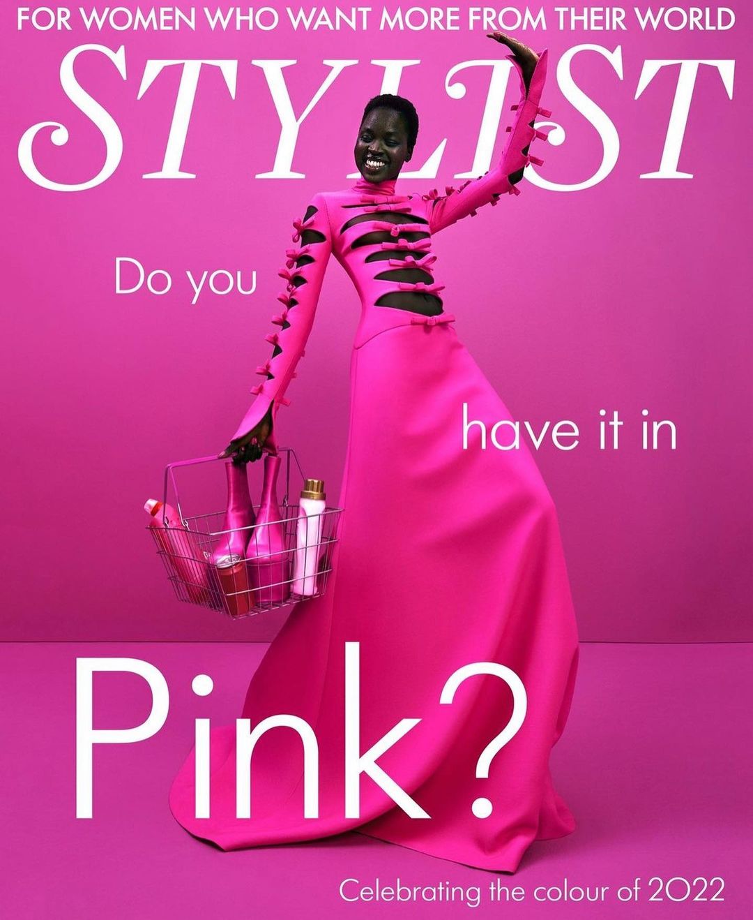 A woman in a vibrant pink gown holds a basket filled with pink products against a matching pink background for Stylist magazine.