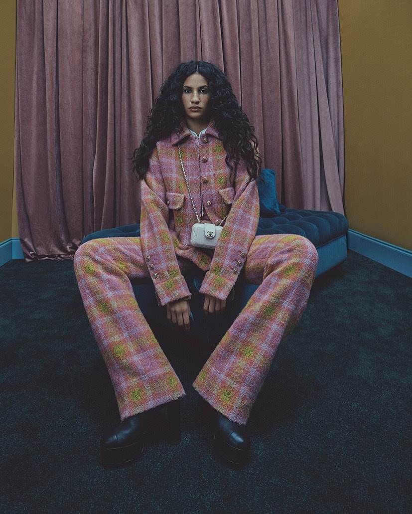 A woman poses indoors wearing a pink plaid tweed suit with a small, white purse. She sits on a dark blue bench with a pink curtain.