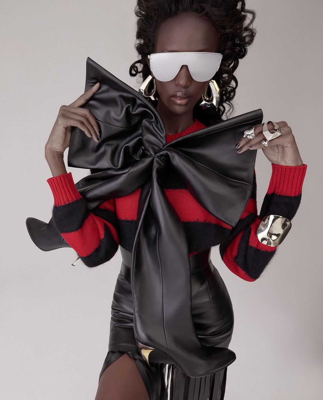 Fashion-forward woman in a striking pose, wearing a leather bow-tie top, skirt, metallic accessories, and futuristic sunglasses against a plain background.