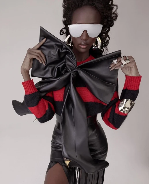 Fashion-forward woman in a striking pose, wearing a leather bow-tie top, skirt, metallic accessories, and futuristic sunglasses against a plain background.
