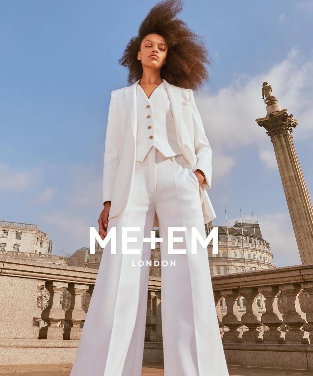 Woman in white suit poses on a city balcony. Tall column with a statue in background. Blue sky with scattered clouds overhead.