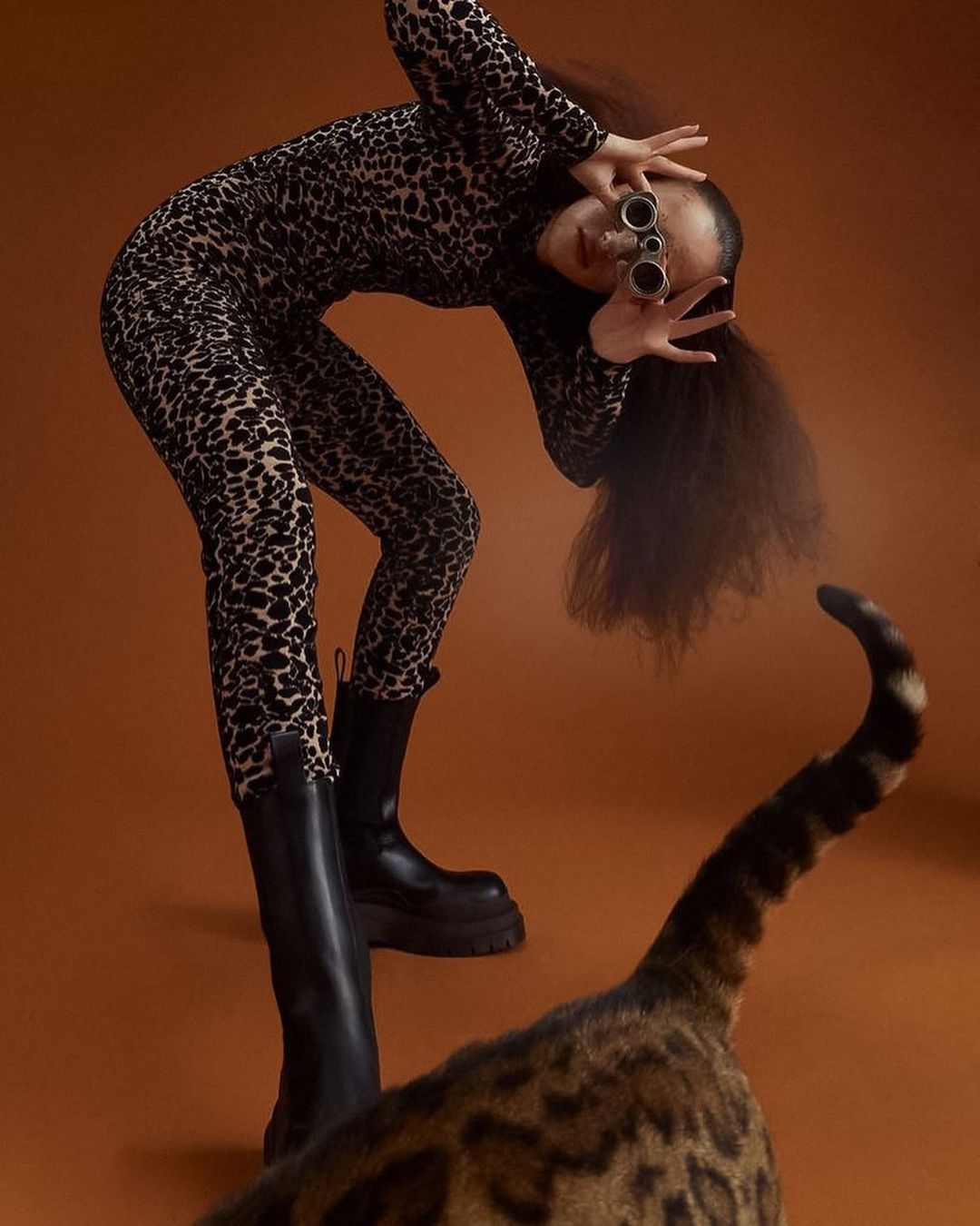 Fashionable woman in leopard print jumpsuit and black platform boots poses with vintage opera glasses and a leopard-patterned cat.