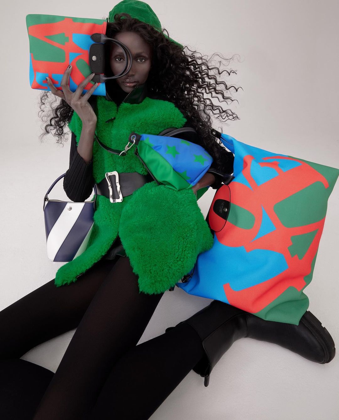 Dark-skinned model poses in green outfit surrounded by colorful handbags. High-fashion editorial shot indoors.