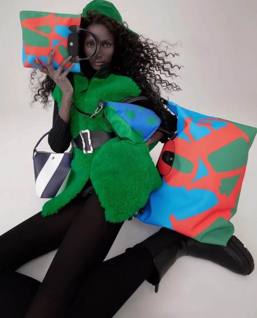 Dark-skinned model poses in green outfit surrounded by colorful handbags. High-fashion editorial shot indoors.