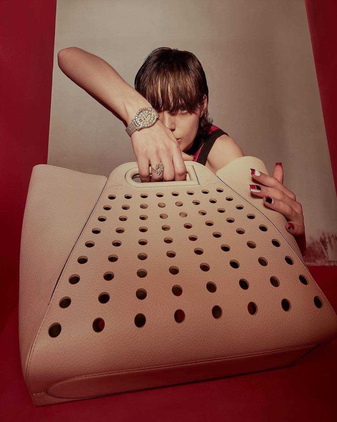 Woman displays a cream-colored tote bag with a circular pattern, wrist watch, and ring against a red and beige background.