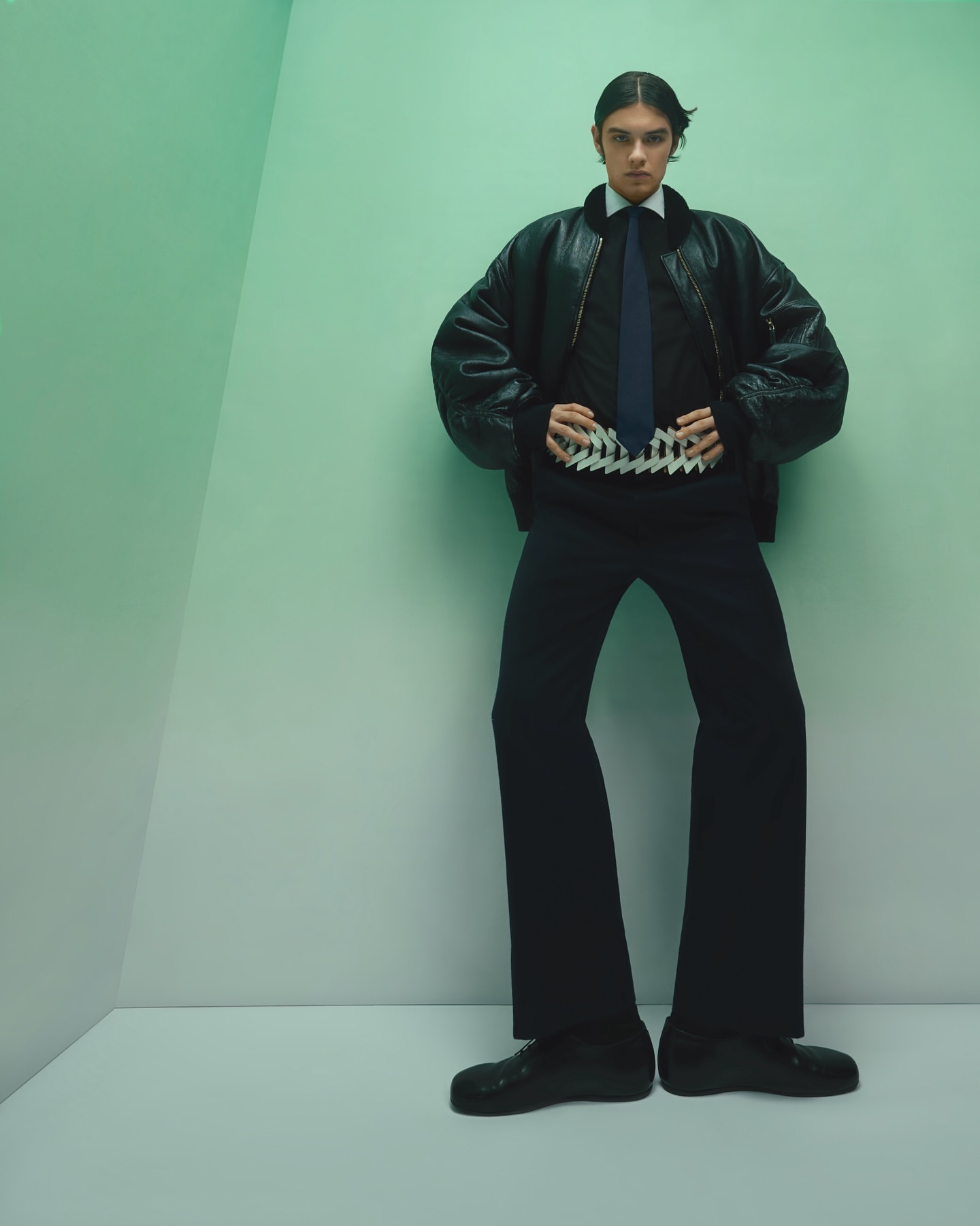 Model wearing a black leather bomber jacket and tie standing against a pale green corner wall