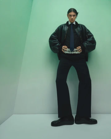 Model wearing a black leather bomber jacket and tie standing against a pale green corner wall