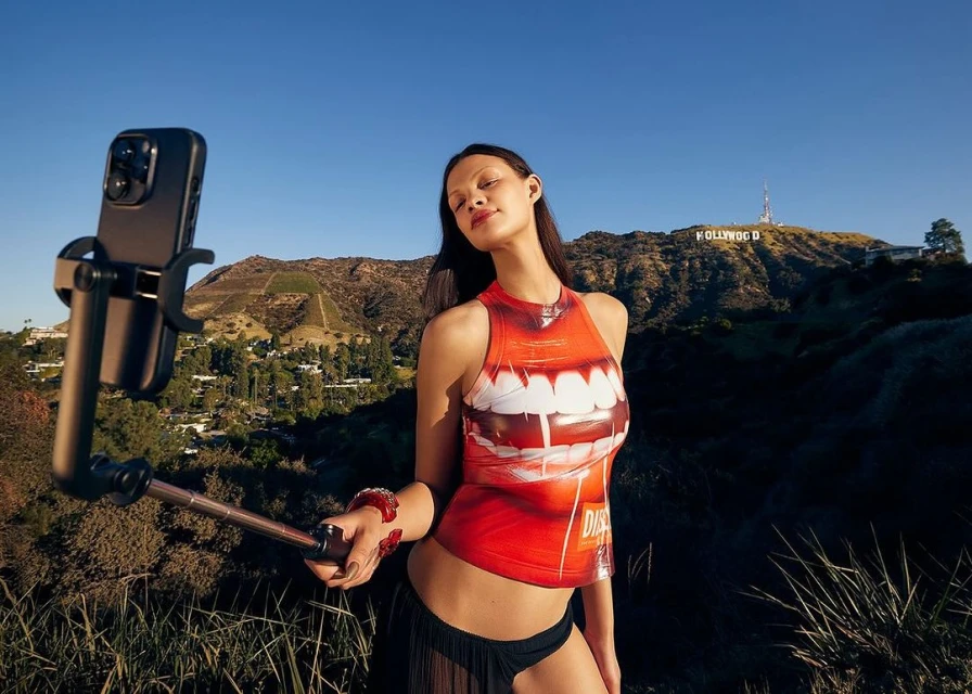 Young woman takes selfie with Hollywood sign visible in background on clear day, wearing orange top with lip graphic design and dark skirt.