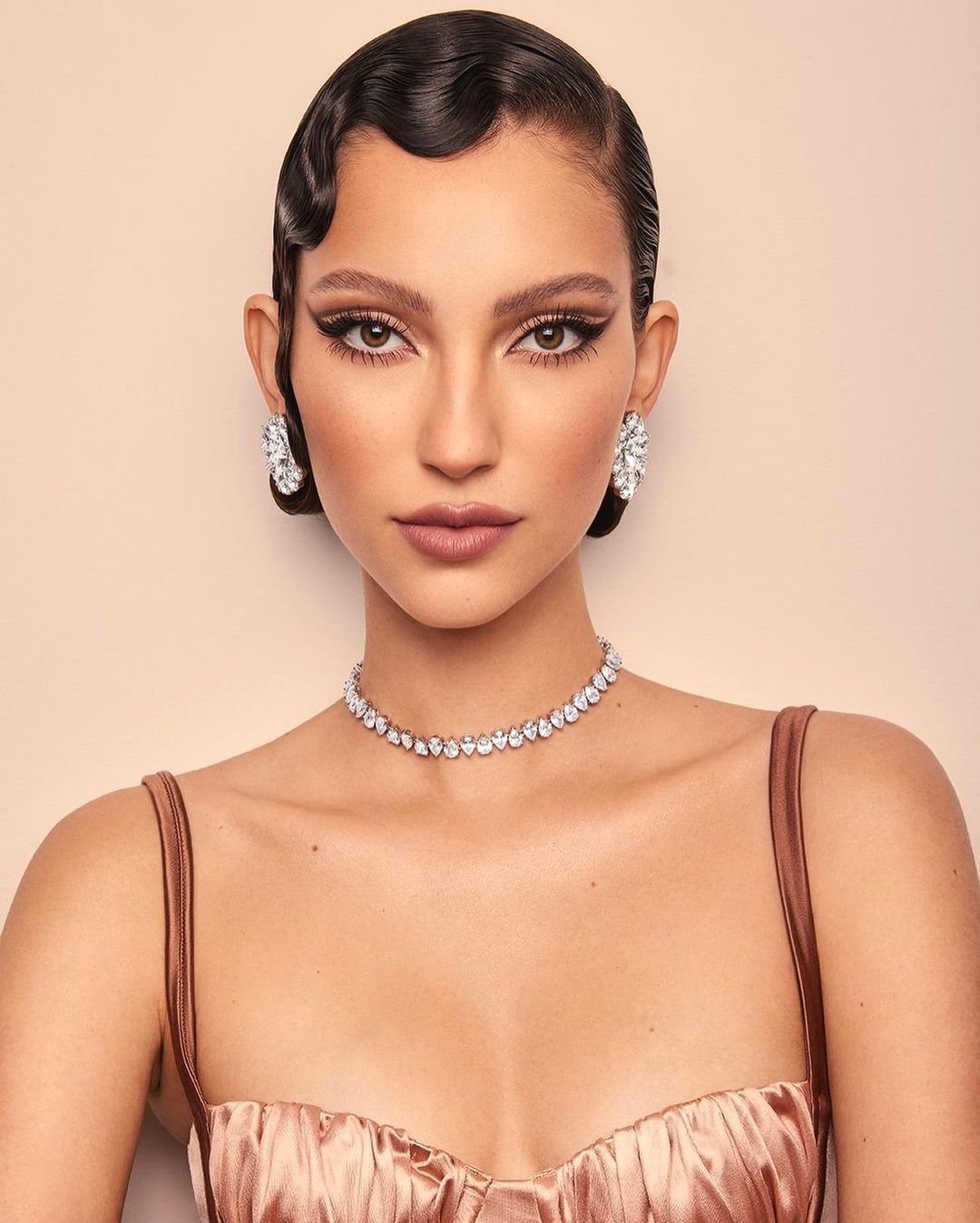 Close-up of a woman with dark hair styled in waves, wearing a champagne-colored dress and diamond jewelry against a neutral background.