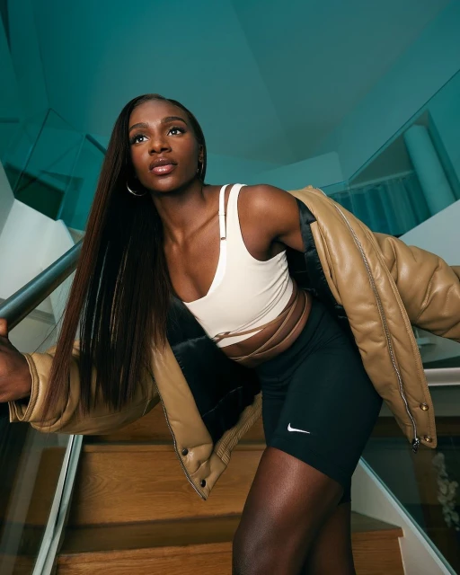 Athletic woman in sports bra and biker shorts poses on stairs with a puffer jacket draped over her shoulder. Indoor setting.