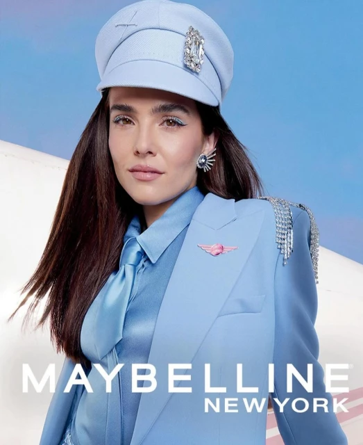 Brunette woman in powder blue suit and cap, commercial studio shot with airplane and sky backdrop, advertising Maybelline New York.