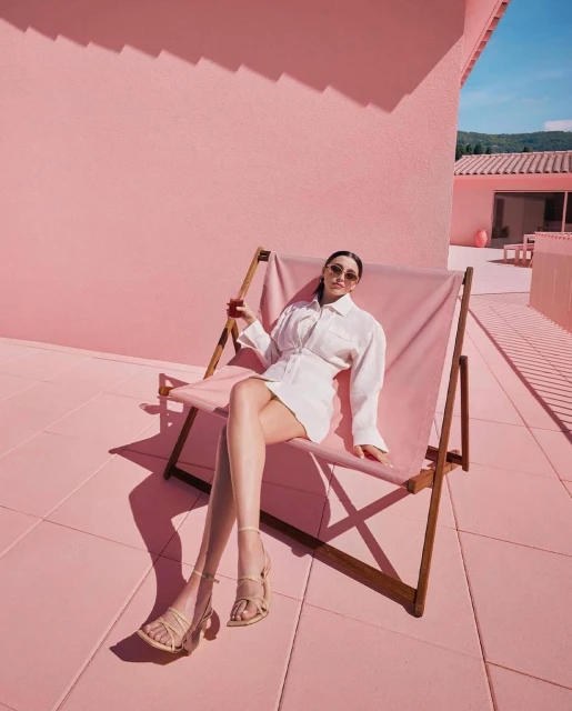 Woman in white dress and sandals seated on a light pink deck chair outdoors, against a light pink stucco wall and tiled floor.