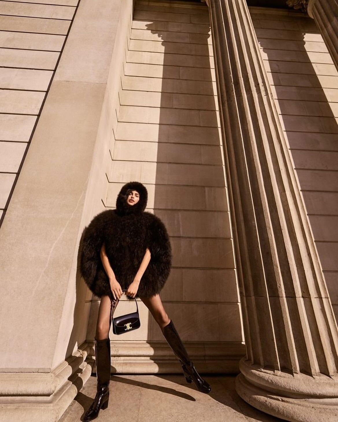 Woman in a fur coat, mini skirt, and boots, standing near stone columns with a black purse. Fashion shot in front of architecture.