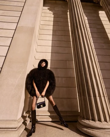 Model in a dark fluffy hooded cape and tall boots holding a black handbag between classical stone columns.