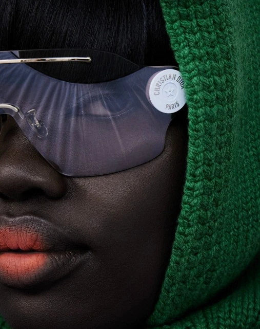 Close-up of a Black woman wearing sunglasses and a green knitted hood. The lighting emphasizes textures and the sunglasses' reflective surface.