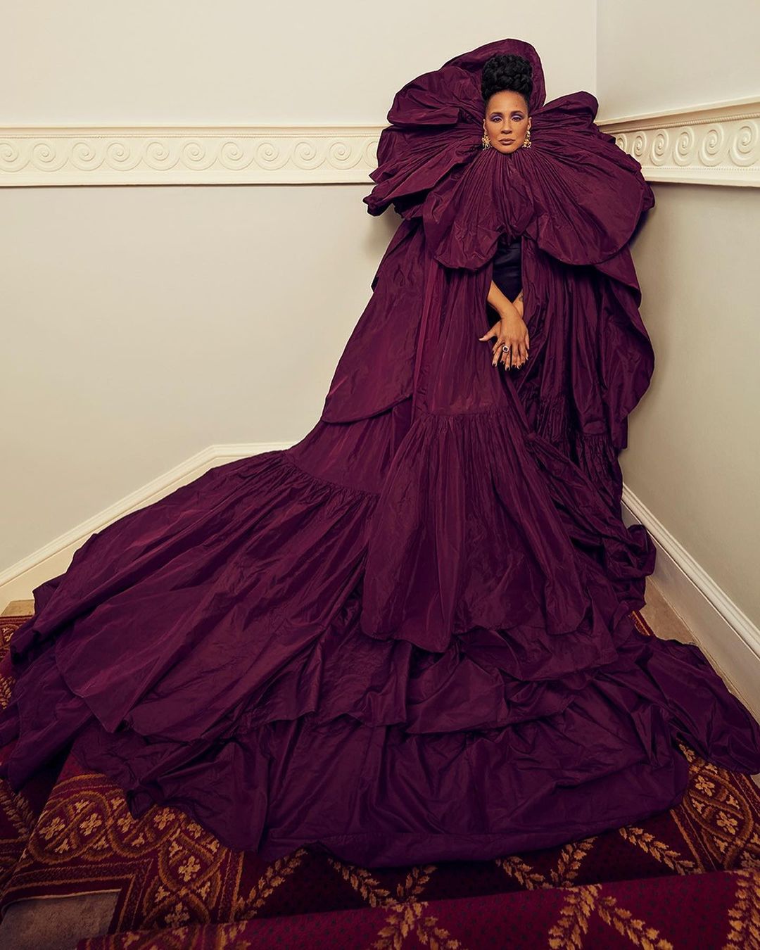 Adult female fashion model poses in a large ruffled burgundy dress with large flower ruff in an indoor setting on a stairwell.