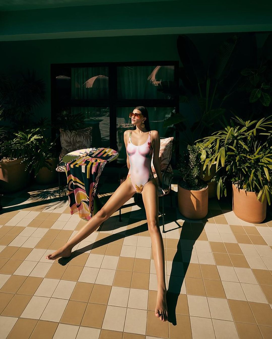 Woman in a patterned swimsuit sits on a chair on a checkered patio surrounded by greenery, bathed in sunlight during daytime.