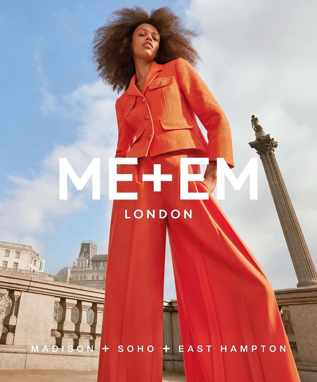 Fashion shot of a Black female model wearing an orange blazer and palazzo pants, posing outdoors against a cloudy blue sky, ME+EM London ad.