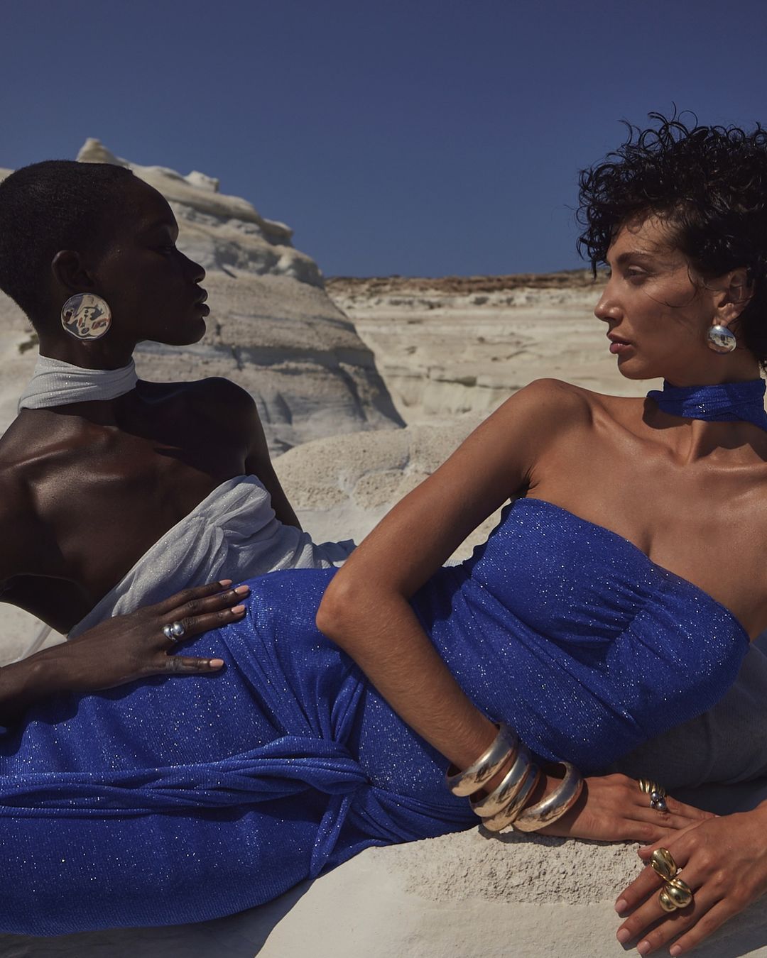 Two women wearing elegant dresses and jewelry pose on a white stone surface with a clear blue sky in the background for a fashion editorial.