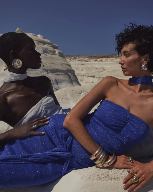 Two women wearing elegant dresses and jewelry pose on a white stone surface with a clear blue sky in the background for a fashion editorial.