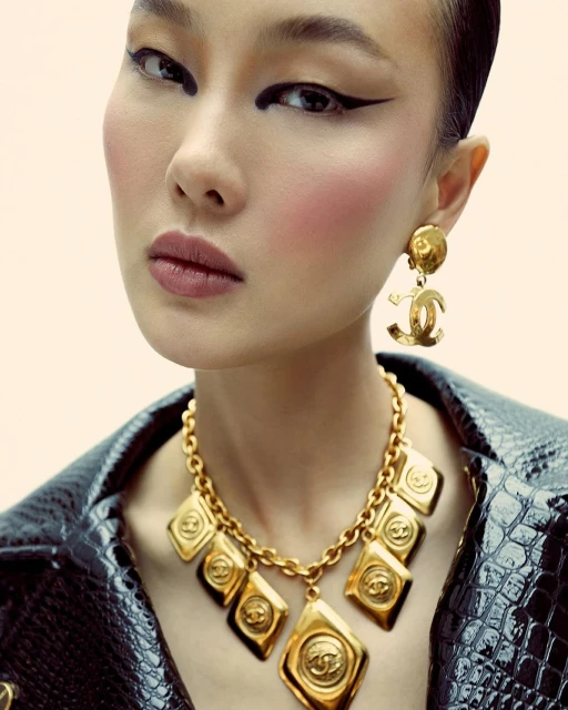 Close-up studio shot of an Asian female model wearing an all-black reptile print jacket with heavy gold Chanel jewelry on light pink background.