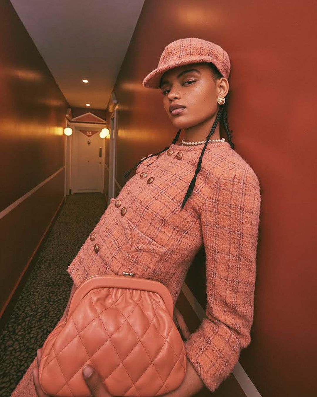 A woman in a peach tweed suit and matching cap stands in a hallway. She holds a quilted clutch bag, looking toward the camera.