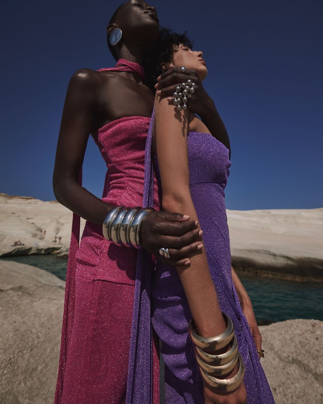 Two women pose in shimmering dresses on a rocky beach under a blue sky, accessorized with bold silver jewelry and metallic bangles.