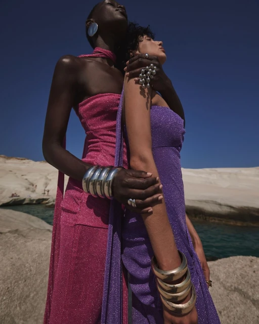 Two women pose in shimmering dresses on a rocky beach under a blue sky, accessorized with bold silver jewelry and metallic bangles.