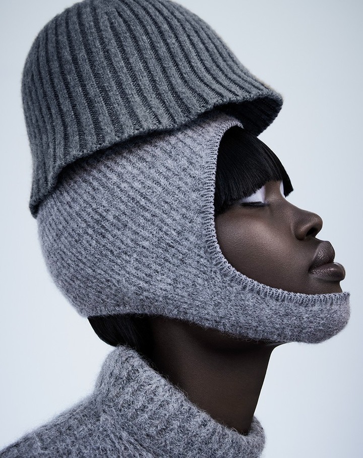 Dark-skinned woman poses in a grey knit balaclava and hat against a light background in a studio shot.