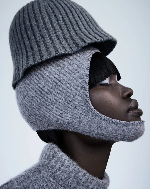Dark-skinned woman poses in a grey knit balaclava and hat against a light background in a studio shot.