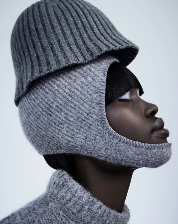 Dark-skinned woman poses in a grey knit balaclava and hat against a light background in a studio shot.