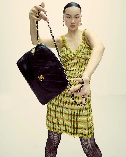 Woman poses indoors holding a large black quilted leather Chanel purse with gold hardware, wearing a yellow plaid dress and fishnet stockings.