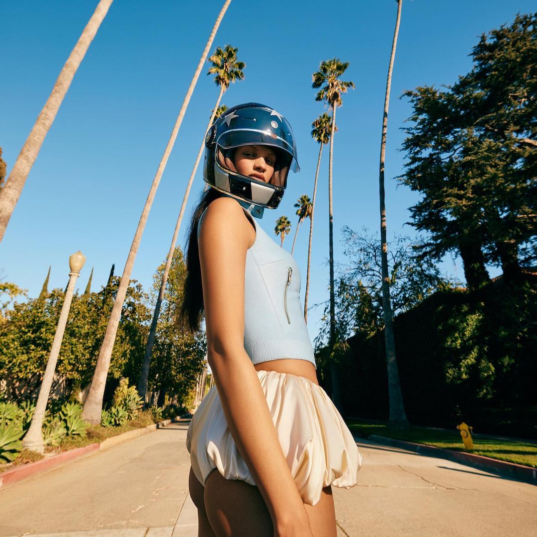 Woman in helmet and pale-yellow shorts stands on a street with palm trees on a sunny day, fashion concept and street photography.