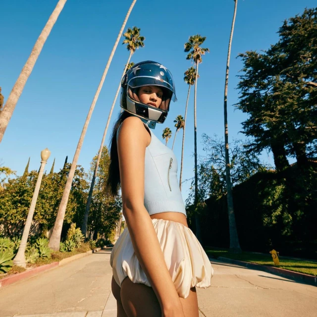 Woman in helmet and pale-yellow shorts stands on a street with palm trees on a sunny day, fashion concept and street photography.