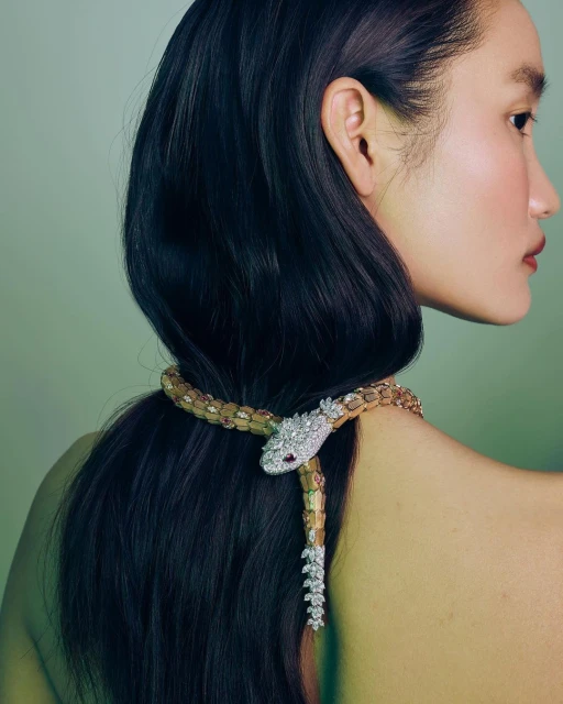 Asian woman with long dark hair poses wearing a gold and diamond snake necklace in a studio setting with soft lighting and green background.