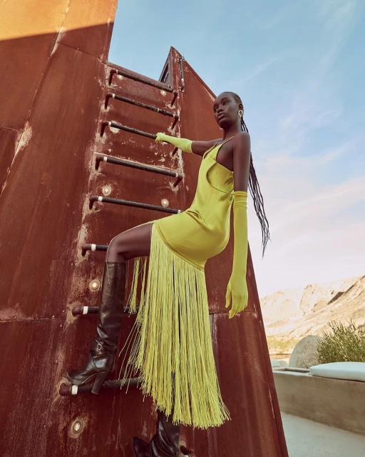 An adult woman models a bright yellow dress and knee-high boots while climbing a rusty metal structure outdoors in a fashion editorial photo.