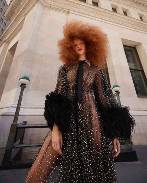 Woman with red afro wears a black embellished gown with feather cuffs standing outdoors against a neutral stone building in daylight.