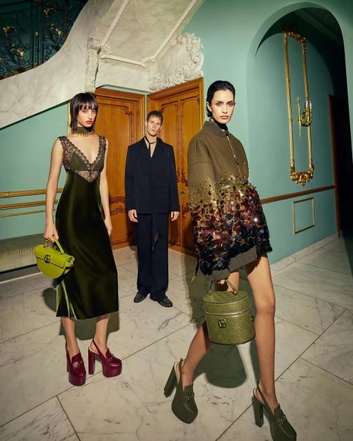 Three fashion models pose indoors. Two women in dresses and heels hold handbags, while a man in a suit stands in the background.