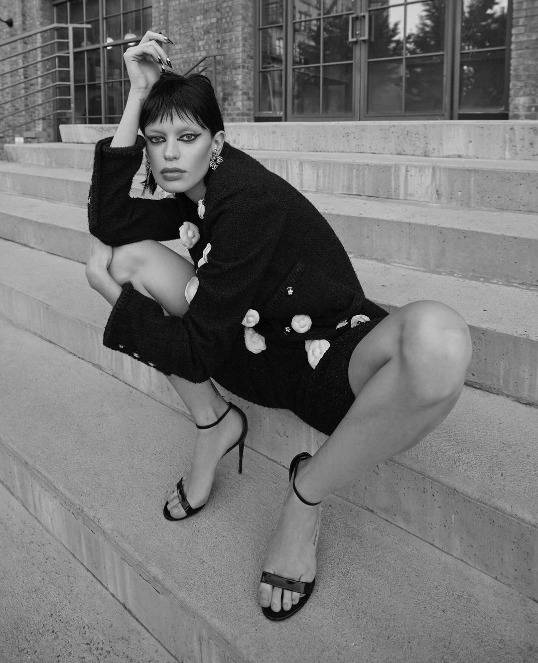 Monochrome portrait of a woman crouching on outdoor concrete stairs wearing a dark textured jacket and heels