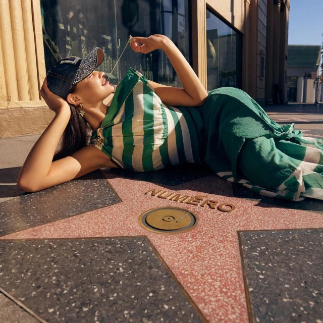 Young woman in a striped dress and baseball cap lying on Hollywood Walk of Fame star with 'Numero' text in outdoor setting.