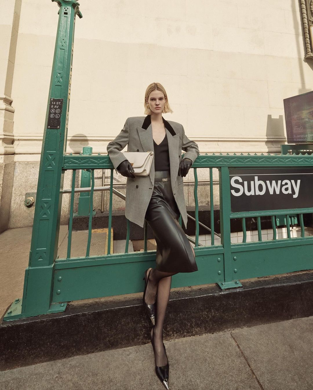 Woman in a blazer and leather skirt posing at a subway entrance, holding a purse and wearing heels and pantyhose, with natural light.