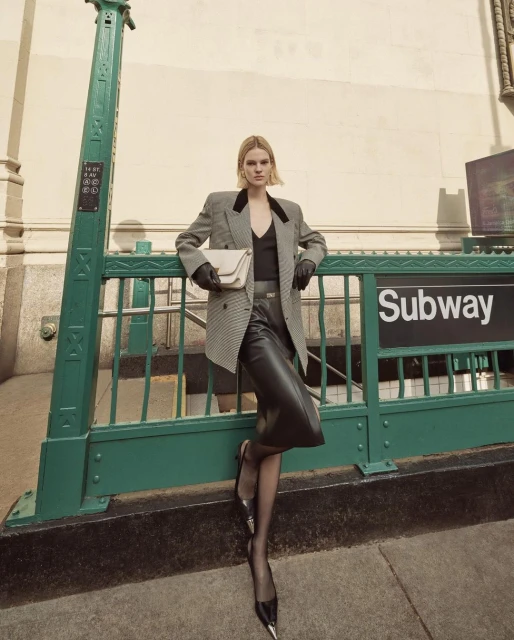 Woman in a blazer and leather skirt posing at a subway entrance, holding a purse and wearing heels and pantyhose, with natural light.