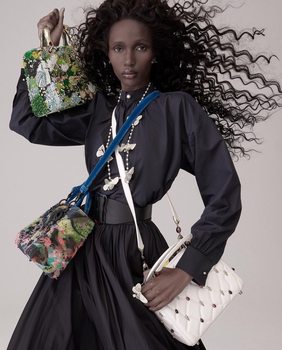 Fashion shot featuring a dark-skinned woman in a black dress, holding and wearing multiple colorful, decorative purses in a studio.