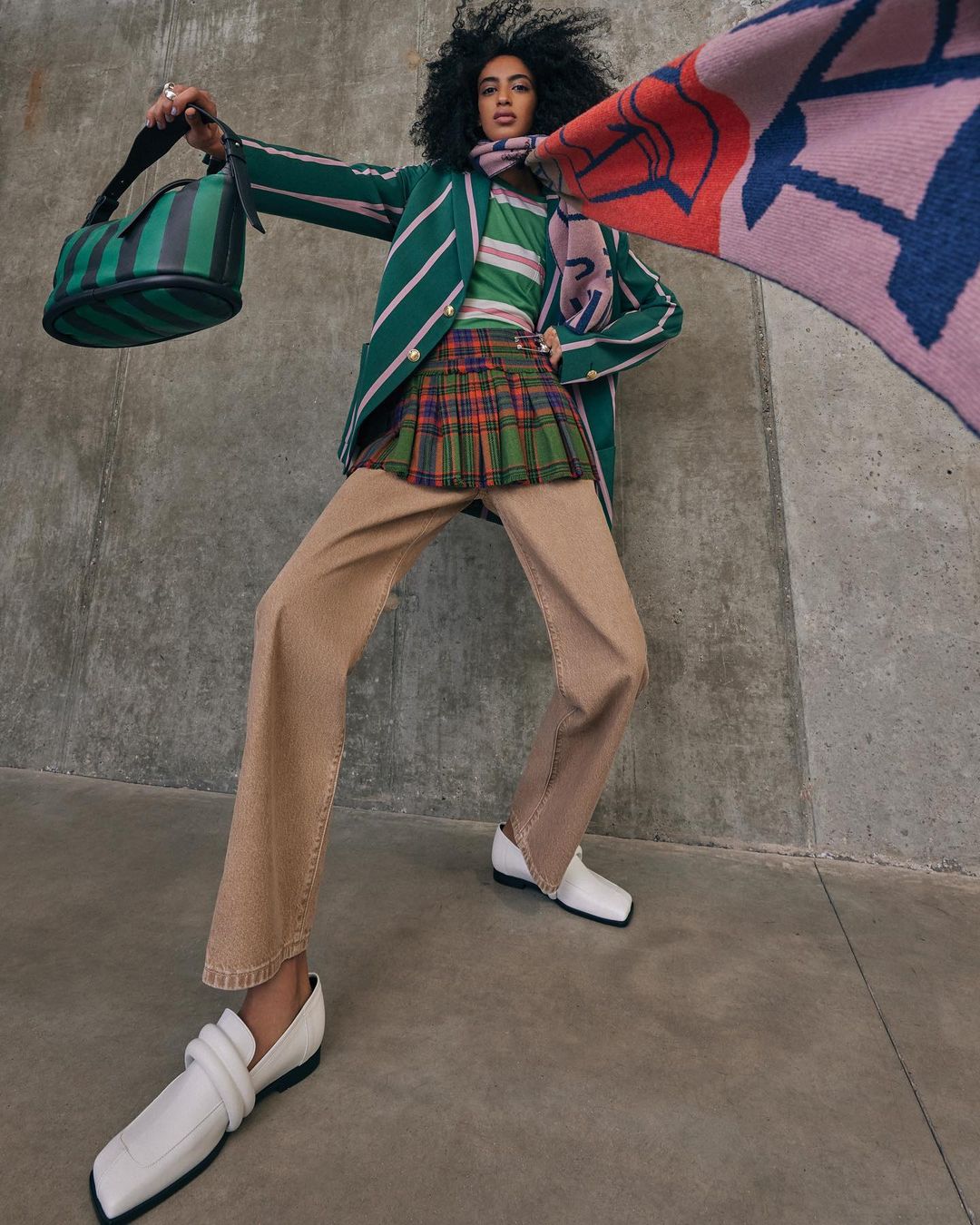 Model with dark curly hair wearing a green striped blazer, plaid miniskirt, and holding a handbag in a low angle shot.
