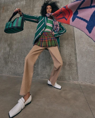 Model with dark curly hair wearing a green striped blazer, plaid miniskirt, and holding a handbag in a low angle shot.