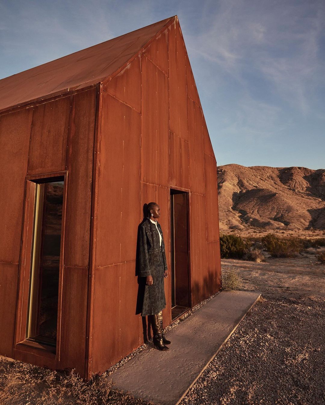 Fashion shot of an adult woman in coat and boots, standing next to a modern cabin in the desert against a clear blue sky.