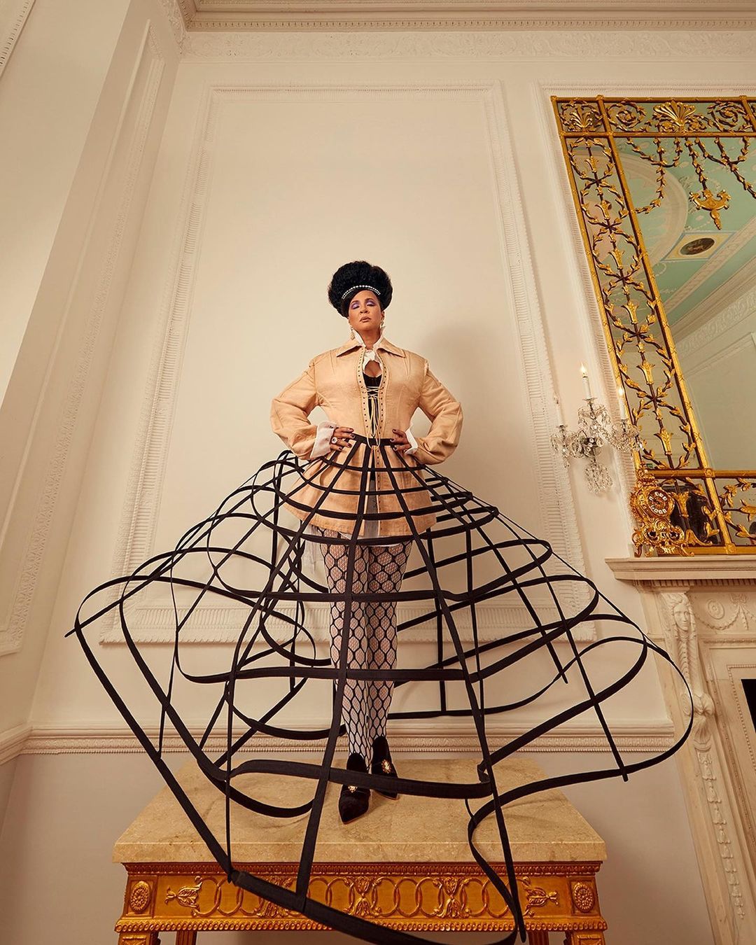 Adult female posing indoors on table with hands on her hips and wearing light peach colored coat with black cage skirt.