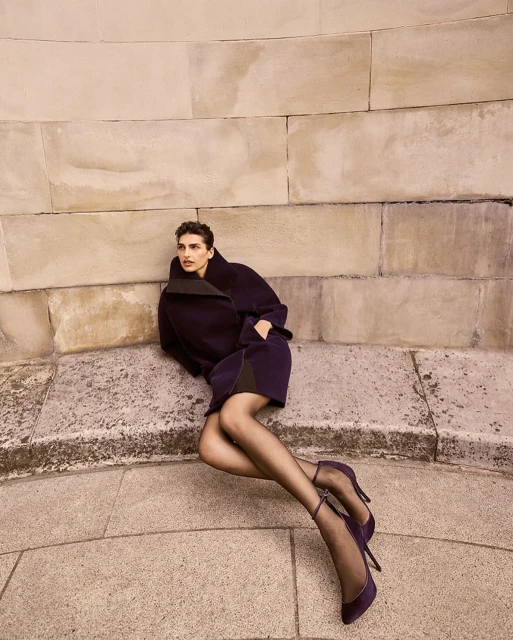 Fashion model wearing a dark coat and heels, sitting on stone steps, posing with a hand in her pocket, looking left.