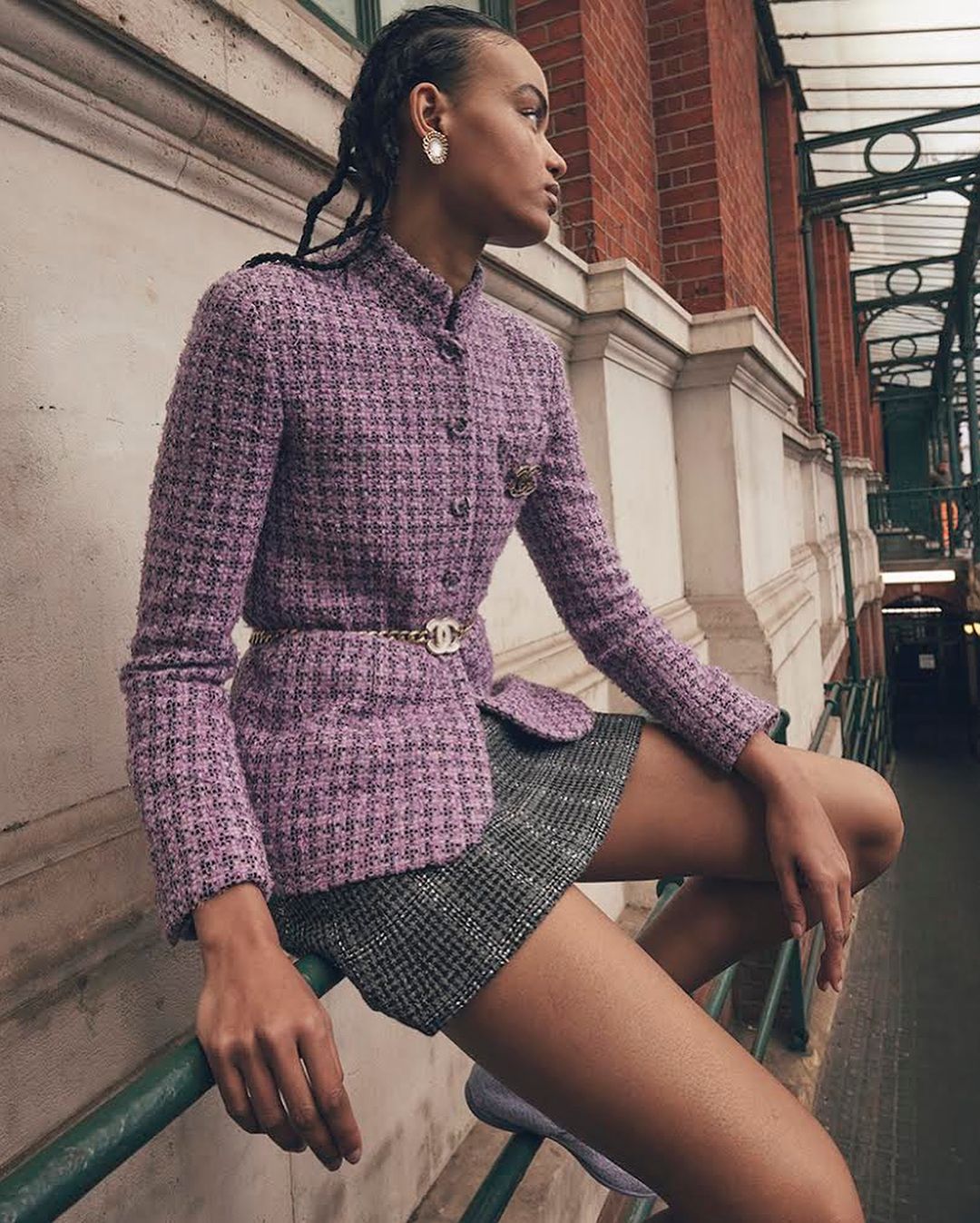 Woman poses wearing lavender tweed jacket and grey plaid shorts outdoors against red brick wall on urban catwalk.