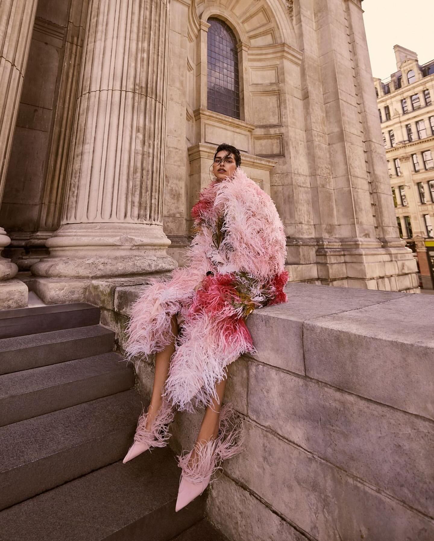 Fashion photography featuring a model in a pink feathered coat and shoes, posing on steps in front of a stone building with pillars.